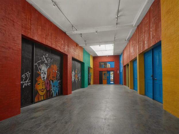 Auf dem Bild ist ein heller, farbenfroher Korridor zu sehen, der von verschiedenen bunten Wänden und Türen geprägt ist. Die Verwendung kräftiger Farben wie Rot, Blau, Grün und Gelb erinnert an die lebendige Straßenkunst und Graffiti-Kultur, die in vielen modernen städtischen Umgebungen, insbesondere in urbanen Zentren, floriert.Diese Art der Gestaltung könnte auch mit kulturellen Initiativen in Verbindung stehen, die darauf abzielen, verlassene oder weniger beachtete Räume zu revitalisieren und mit künstlerischem Ausdruck zu füllen. Solche Projekte fördern oft das Gemeinschaftsgefühl und ermöglichen es Künstlern, ihre Werke einem breiten Publikum zugänglich zu machen.Ein Beispiel für ähnliche kulturelle Initiativen wären die urbanen Kunstbewegungen in Städten wie Berlin und Barcelona, wo Graffiti und Wandmalereien oft Teil der Stadtlandschaft sind und soziale Themen ansprechen. Die Mischung aus Farbe und Kunst in diesem Korridor könnte daher als eine Hommage an das kreative Potential in städtischen Räumen interpretiert werden, das gleichzeitig auch historische und gesellschaftliche Kontexte reflektiert.
