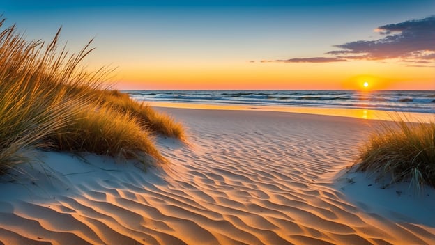 Ein Strand mit feinem Sand, Dünen und Gras im Vordergrund, während die Sonne am Horizont untergeht und das Wasser sanft wellt.